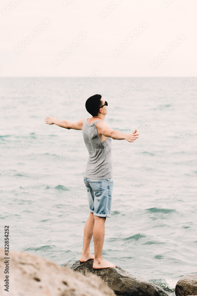 a young guy in a t shirt and shorts with his arms outstretched stands on the beach on large rocks