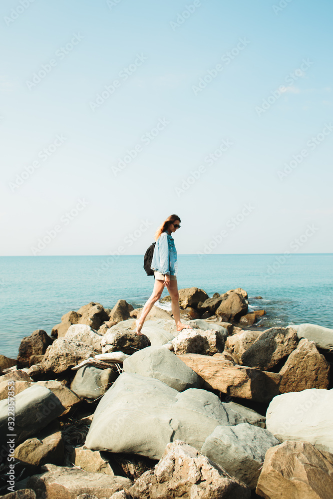 a young girl traveler with a backpack walks barefoot on large stones on the sea coast a young girl traveler with a backpack walks barefoot on large stones on the sea coast