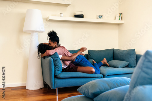 Joyful black young girlfriends hugging on couch at home