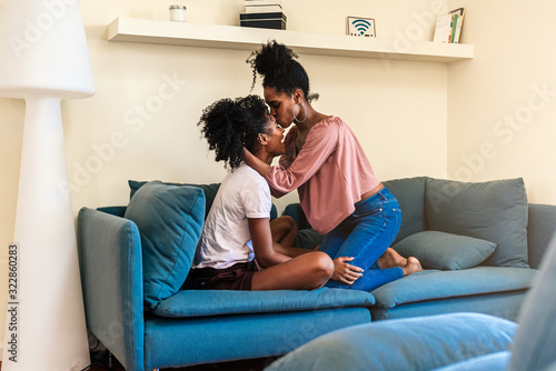Joyful black young girlfriends hugging on couch at home