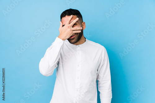 Young latin man against a blue background isolated blink at the camera through fingers, embarrassed covering face.