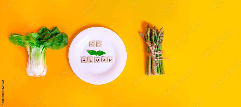 Top view Pak Choi, asparagus and white plate with Go vegan message on wooden blocks on orange background. Vegetarian and vegan diet. Veganism. Sustainable lifestyle, plant-based foods. Wide banner.
