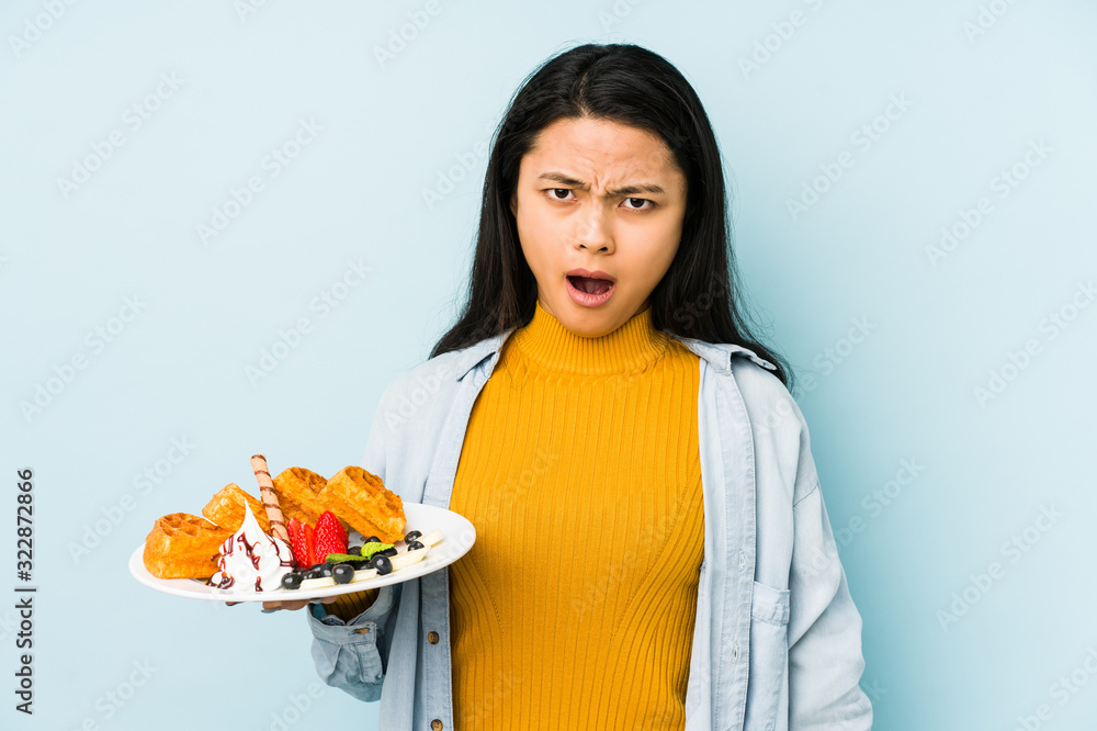 Young chinese woman holding waffle isolated on blue background joyful and carefree showing a peace symbol with fingers. Young chinese woman holding waffle isolated on blue background joyful and carefree showing a peace symbol with fingers.