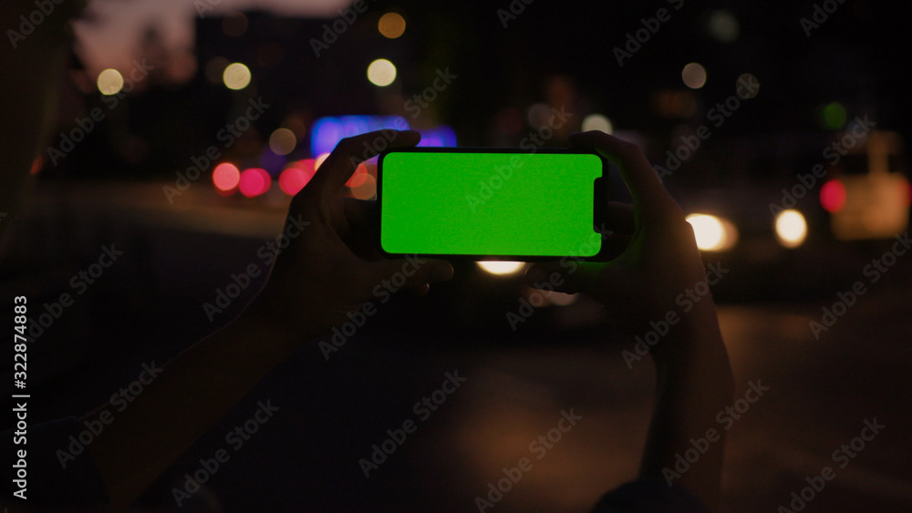Lviv, Ukraine - May 19, 2018: Handheld shot of young woman holding horizontal smartphone watching green screen mock-up device standing at traffic road in the evening. Urban life and technology.
