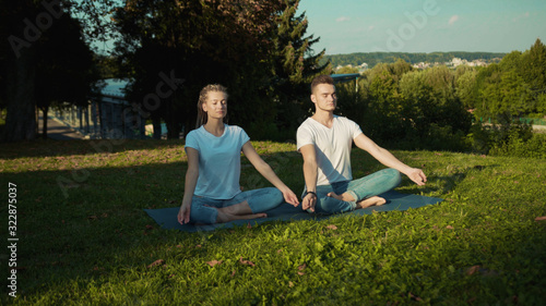 Young woman training in yoga class with handsome instructor sitting on grass breathing relaxing on fresh summer morning in the park.