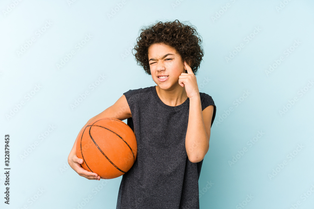 Kid boy playing basketball isolated on blue background covering ears with hands.