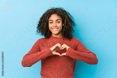 Young african american curly hair woman smiling and showing a heart shape with hands.