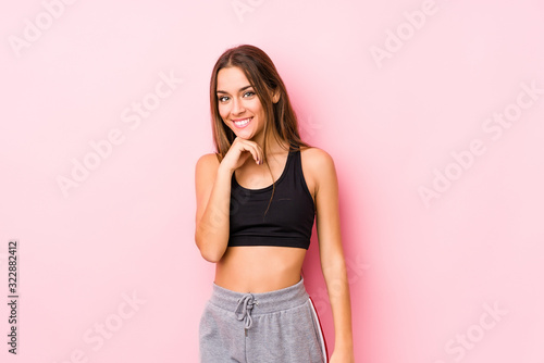 Young caucasian fitness woman posing in a pink background smiling happy and confident  touching chin with hand.