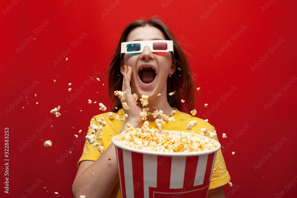 funny young girl shocked in 3D glasses watching a movie and eating popcorn on a red colored background, she screams and popcorn flies