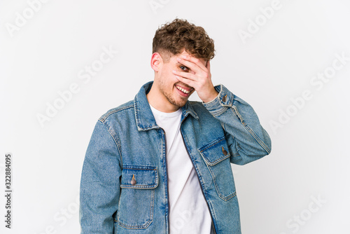 Young blond curly hair caucasian man isolated blink at the camera through fingers, embarrassed covering face.