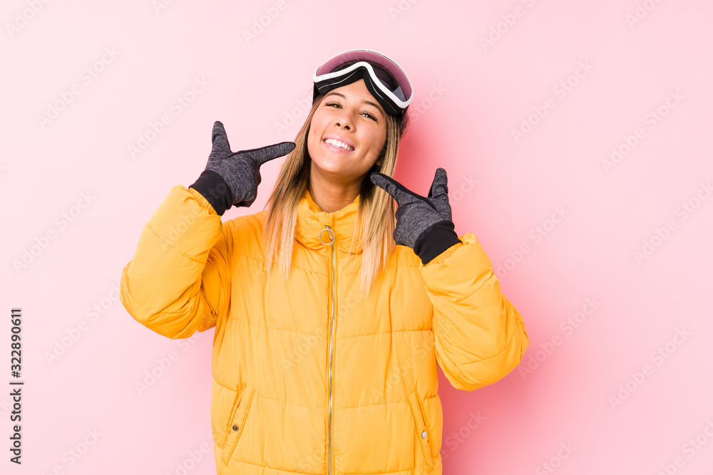 Young caucasian woman wearing a ski clothes in a pink background smiles, pointing fingers at mouth. Young caucasian woman wearing a ski clothes in a pink background smiles, pointing fingers at mouth.
