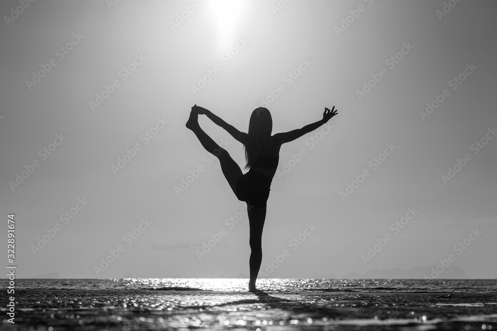 Fototapeta premium Silhouette of woman standing at yoga pose on the tropical beach during sunset. Girl practicing yoga near sea water. Black and white
