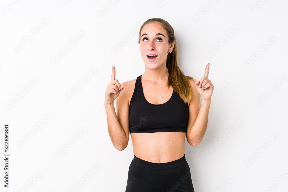 Young caucasian fitness woman posing in a white background pointing upside with opened mouth. Young caucasian fitness woman posing in a white background pointing upside with opened mouth.