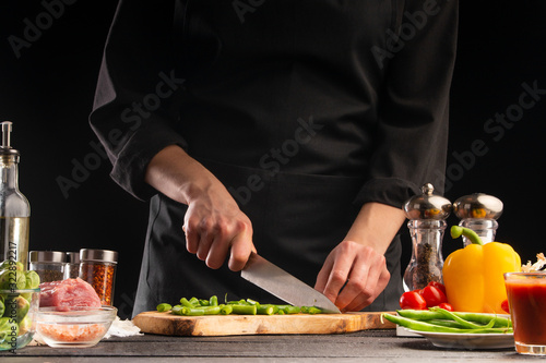 The chef cuts green beans on the background of vegetable ingredients, on a black background. Cooking salad. Healthy and wholesome food, cuisine and cooking, recipe book.