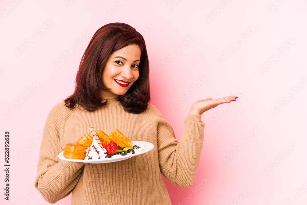 Middle age latin woman holding a waffle isolated showing a copy space on a palm and holding another hand on waist. Middle age latin woman holding a waffle isolated showing a copy space on a palm and holding another hand on waist.