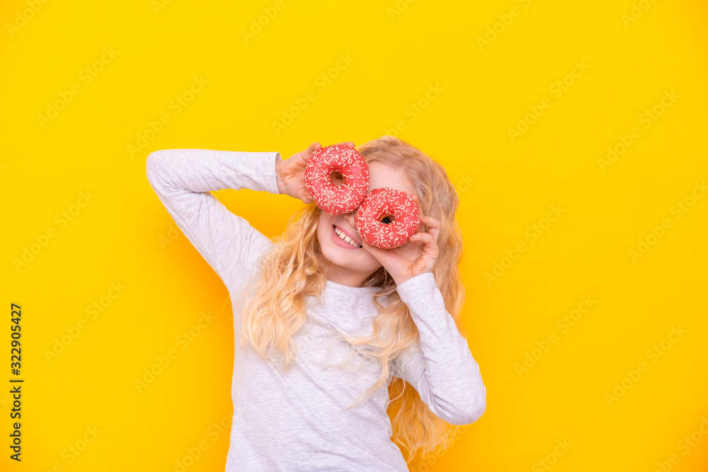 Crazy cheerful blonde girl in birthday hat smiling, having fun and looking through two red donuts on her eyes. Sweets. Yellow studio background.