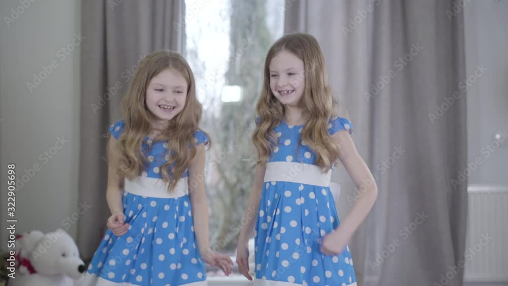 Middle shot of joyful twin sisters having fun indoors. Joyful brunette Caucasian girls in similar blue dotted dresses grimacing and smiling. Leisure, fun, lifestyle, family. Middle shot of joyful twin sisters having fun indoors. Joyful brunette Caucasian girls in similar blue dotted dresses grimacing and smiling. Leisure, fun, lifestyle, family.