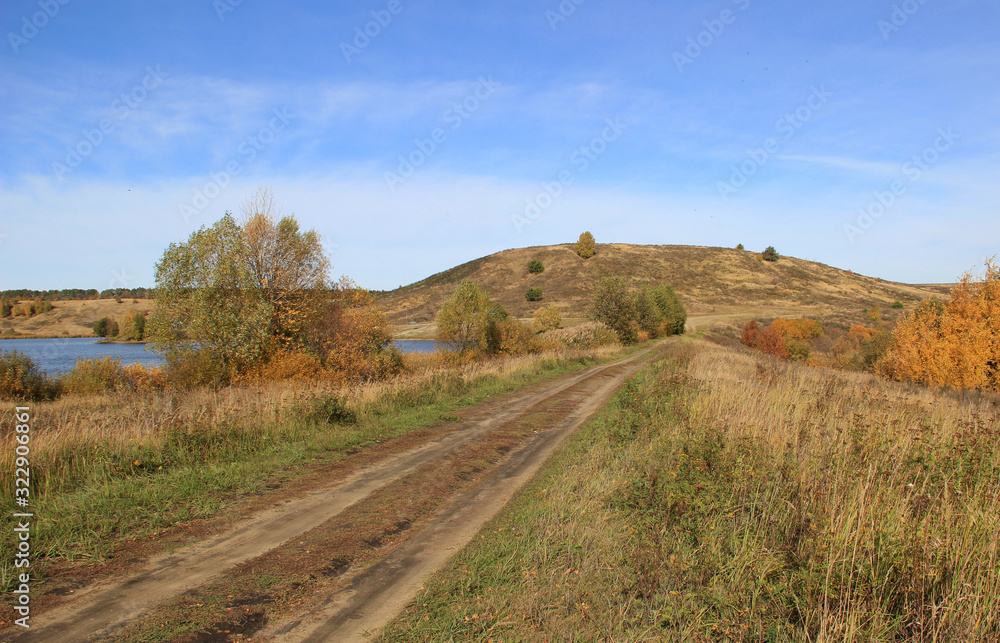 A country road follows the dam to a small hill. On one side is a pond against the blue sky.