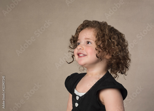Curly hair adorable happy toddler girl smiling looking away from camera isolated on brown background