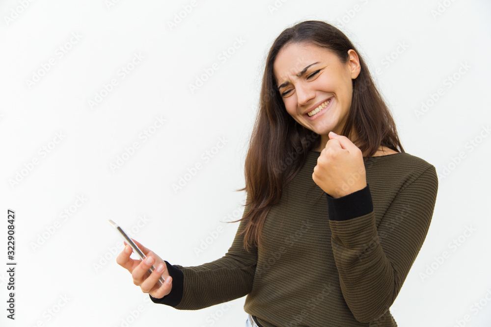 Successful young woman with raised fist looking at phone. Smiling lady holding modern phone. Technology, success concept Successful young woman with raised fist looking at phone. Smiling lady holding modern phone. Technology, success concept