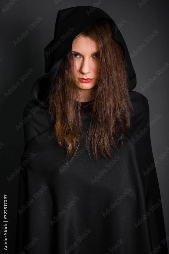 Fototapeta premium Young, beautiful girl in a black cloak with a hood in the studio on a gray background. Long hair fluttering on the wind. A model with clean skin. Suit of a fortuneteller, witches for Halloween.