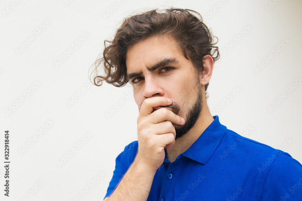 Serious young man holding hand on mouth. Thoughtful bearded guy looking at camera. Thinking, doubting concept Serious young man holding hand on mouth. Thoughtful bearded guy looking at camera. Thinking, doubting concept