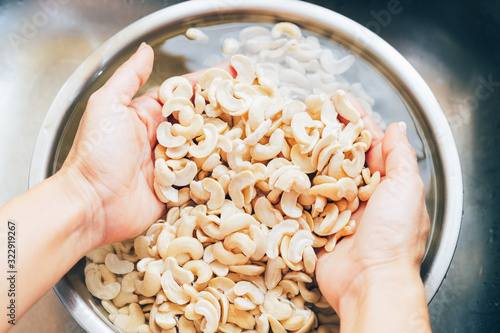 Fotografie Closeup, woman’s hands wash, scrub, soak and scoop cashew nuts from water in bowl to remove dirts, tannins, phytic acid on surface, healthy & good for digestion of plant based diet, vegan, vegetarian