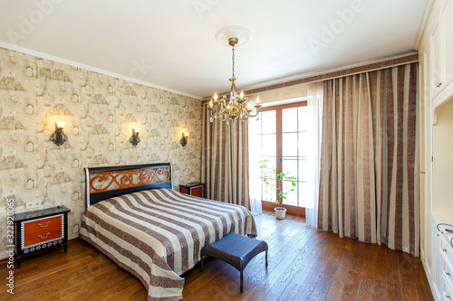 Elegant expensive bedroom with a large white bed with a striped bedspread. A high headboard with a red pattern, bedside tables with red drawers, Twisted wall lamps and a chandelier. 