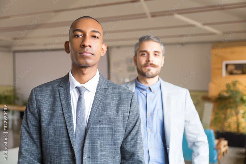Two confident businessmen looking at camera. Concentrated young men wearing formal wear posing in office. Business, confidence concept Two confident businessmen looking at camera. Concentrated young men wearing formal wear posing in office. Business, confidence concept