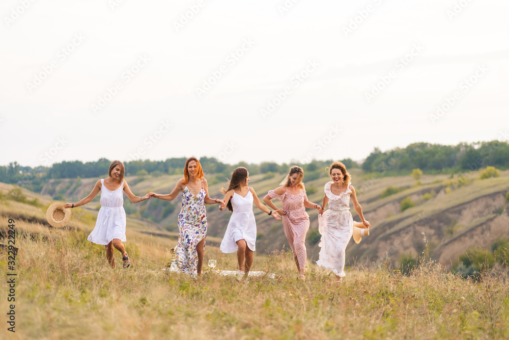 Obraz premium The company of cheerful female friends have a great time together on a picnic in a picturesque place overlooking the green hills. Girls in white dresses dancing in the field