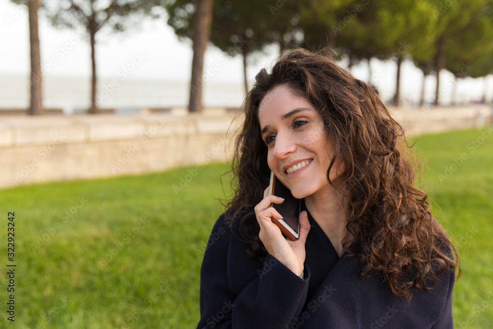 Smiling woman talking on smartphone in park. Cheerful curly lady wearing coat communicating via phone. Technology, communication concept Smiling woman talking on smartphone in park. Cheerful curly lady wearing coat communicating via phone. Technology, communication concept