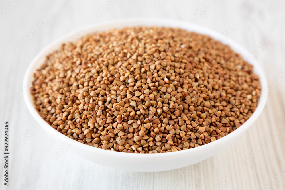 Uncooked Roasted Buckwheat in a white bowl on a white wooden surface, side view. Close-up. Uncooked Roasted Buckwheat in a white bowl on a white wooden surface, side view. Close-up.