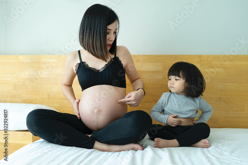 Mother and daughter sit on bed showing  tummy.