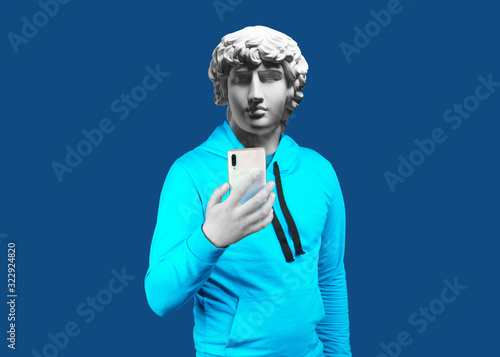 Modern art collage. Concept portrait of a man  holding mobile smartphone using app texting sms message. Gypsum head of Antinous. Man in suit. On a blue background. photo