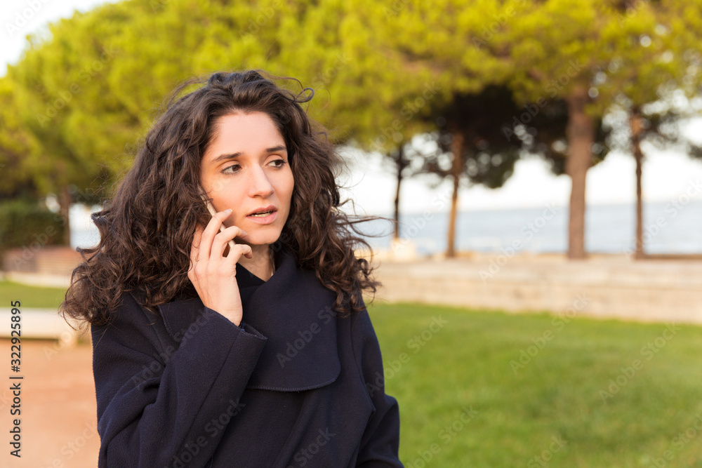 Thoughtful woman talking on smartphone while walking on street. Serious curly lady wearing coat communicating via phone. Technology, communication concept Thoughtful woman talking on smartphone while walking on street. Serious curly lady wearing coat communicating via phone. Technology, communication concept