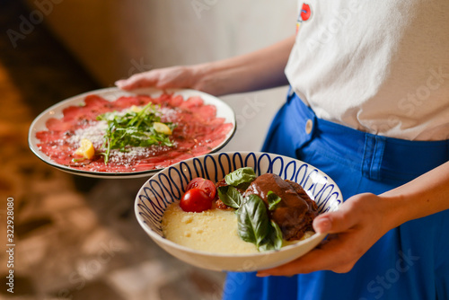 Carpaccio of beef served on a white plate. Waiter holds two plates with Italian food in Italian restaurant.