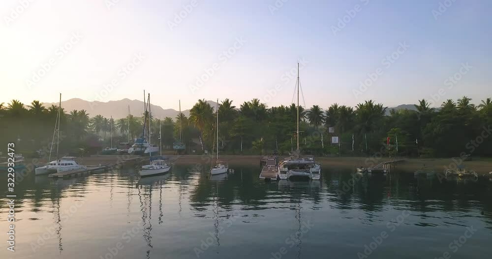 Sailboats in sunrise with palm trees and mountains in far back. Calm morning aerial shot flying up and backwards from a bay in Lombok, Indonesia. Sailboats in sunrise with palm trees and mountains in far back. Calm morning aerial shot flying up and backwards from a bay in Lombok, Indonesia.