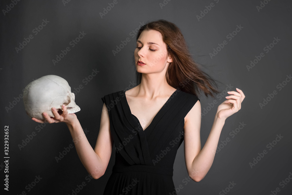 Naklejka premium A beautiful, young girl with long hair in a black dress with a skull in her hands. Studio photo on a gray background. Witchcraft, necromancy, fortune telling, Halloween.