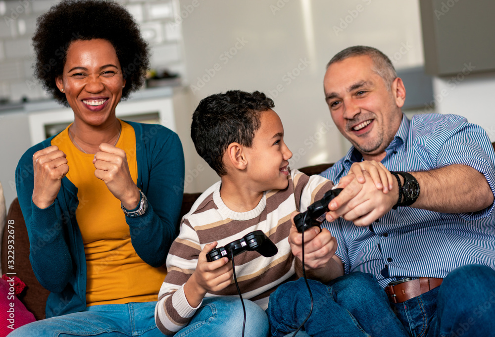 Smiling mixed race family enjoying time at home sitting on sofa in living room and playing video games. - obrazy, fototapety, plakaty 