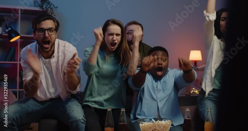Excited group of people rejoicing together while watching sport game at home. Happy friends sitting in front of tv on sofa, supporting and looking overjoyed by moment . Concept of leisure.
