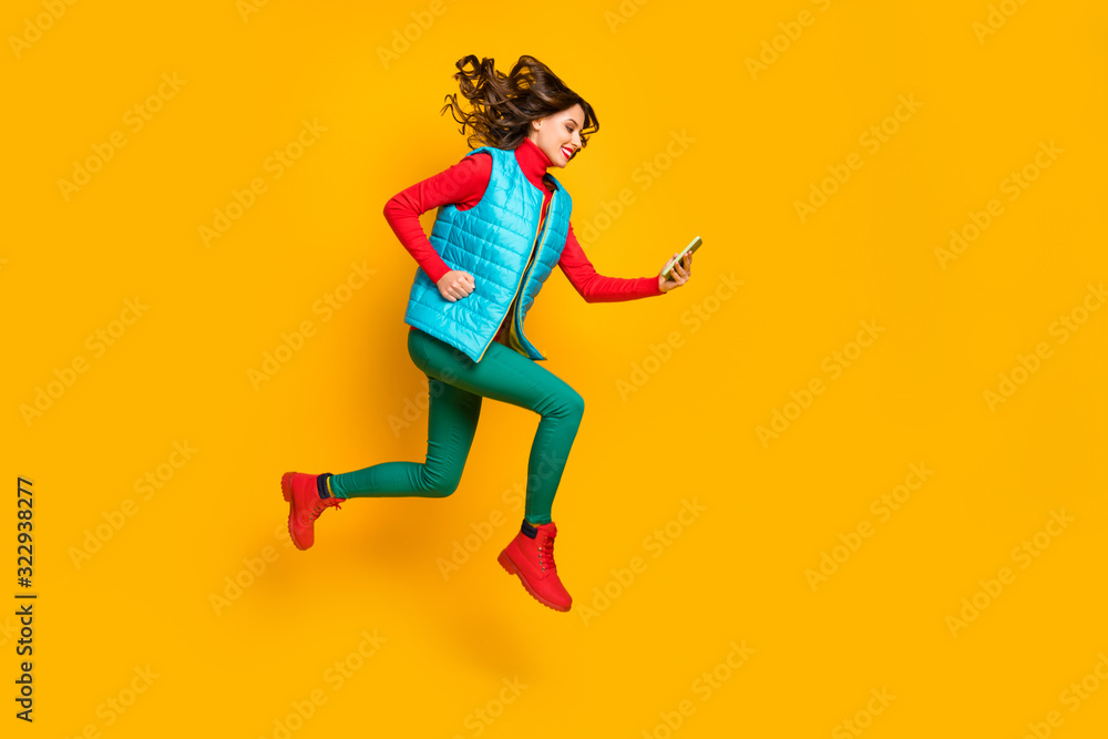 Full size profile side photo cheerful girl student jump use smartphone follow social media ads run hurry wear blue vest red green pants trousers shoes isolated bright yellow color background