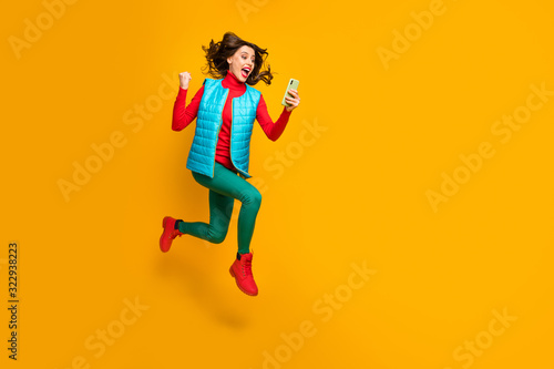 Full size photo delighted girl use smartphone follow social media autumn discount win lottery raise fists scream jump run wear green blue red pants shoes vest isolated yellow color background © deagreez