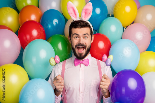 Photo of funny guy colorful decorations easter party festive mood hold eggs on sticks wear pink shirt bow tie suspenders fluffy bunny ears on balloons creative design background