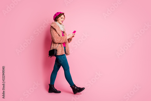 Full size profile photo of pretty lady hold telephone chatting friends walking down street wear casual stylish fur coat scarf beret trousers shoes outfit isolated pink color background
