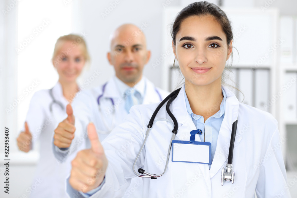Doctor woman showing OK in clinic with colleagues at the background. Hispanic or latin american staff in medicine Doctor woman showing OK in clinic with colleagues at the background. Hispanic or latin american staff in medicine
