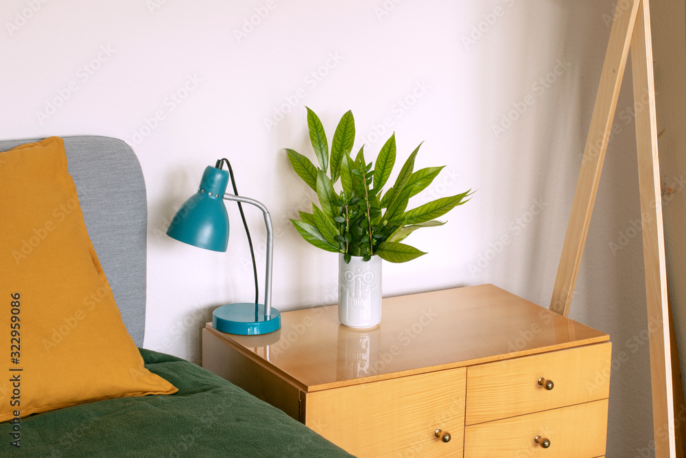 Detail of home interior with turquoise lamp and green leaves decoration standing on wooden sideboard next to bed. Bouquet of fresh leaves indoors. Simple green fresh home floral decoration.