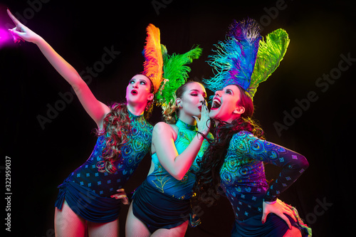 Feelings. Beautiful young women in carnival, stylish masquerade costume with feathers on black background in neon light. Copyspace for ad. Holidays celebration, dancing, fashion. Festive time, party.
