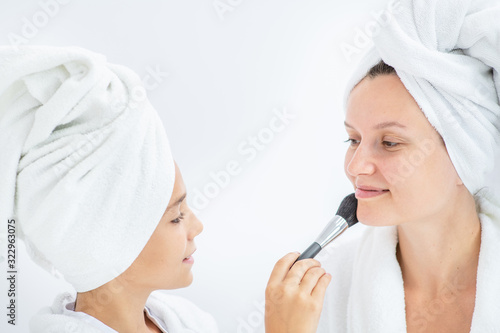 Young girl applying powder on face her mother using a brush. Mom and child girl are in bathrobes and with towels on their heads