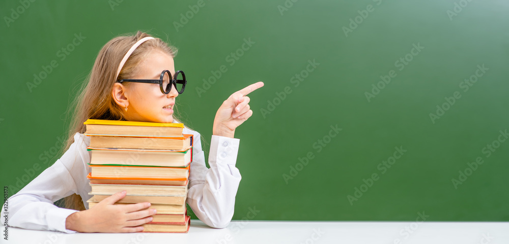 Smart girl wearing eyeglasses sits with books and points away on empty green chalkboard. Empty space for text