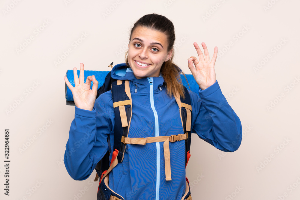 Young mountaineer girl with a big backpack over isolated background showing an ok sign with fingers Young mountaineer girl with a big backpack over isolated background showing an ok sign with fingers